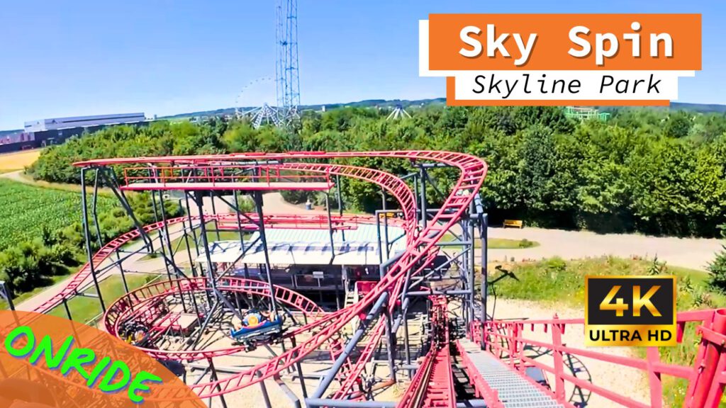 Blick auf die Achterbahn Sky Spin im Skyline Park. Die Bahn besteht aus roten Schienen und schwarzen Stützen, schlängelt sich in engen Kurven und Höhen über den Park, während im Hintergrund grüne Bäume und andere Fahrgeschäfte zu sehen sind. Ein Schild zeigt die Aufschrift "Onride" und das Video ist in 4K Ultra HD verfügbar.