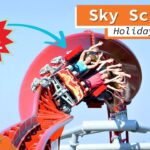 Eine Achterbahn mit dem Namen Sky Scream im Holiday Park führt Fahrgäste durch einen engen Looping. Die Insassen heben die Arme und wirken angespannt, während die Bahn eine kurvenreiche, steile Strecke entlangfährt. Im Hintergrund ist der blaue Himmel zu sehen, und im Bild befinden sich farbenfrohe Grafiken mit den Aufschriften "You are here", "Onride" und "4K Ultra HD".