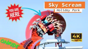 Eine Achterbahn mit dem Namen Sky Scream im Holiday Park führt Fahrgäste durch einen engen Looping. Die Insassen heben die Arme und wirken angespannt, während die Bahn eine kurvenreiche, steile Strecke entlangfährt. Im Hintergrund ist der blaue Himmel zu sehen, und im Bild befinden sich farbenfrohe Grafiken mit den Aufschriften "You are here", "Onride" und "4K Ultra HD".