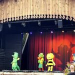 Auf einer hölzernen Freilichtbühne im Holiday Park führen mehrere kostümierte Charaktere eine Kinder-Show auf. Zu sehen sind bekannte Figuren wie ein grüner Drache, ein Papagei, eine gelbe Biene und eine Figur im Wikinger-Kostüm. Der rote Bühnenvorhang und bunte Lichter im Hintergrund schaffen eine lebhafte Atmosphäre. Einige Zuschauer beobachten die Show aus dem Publikum.