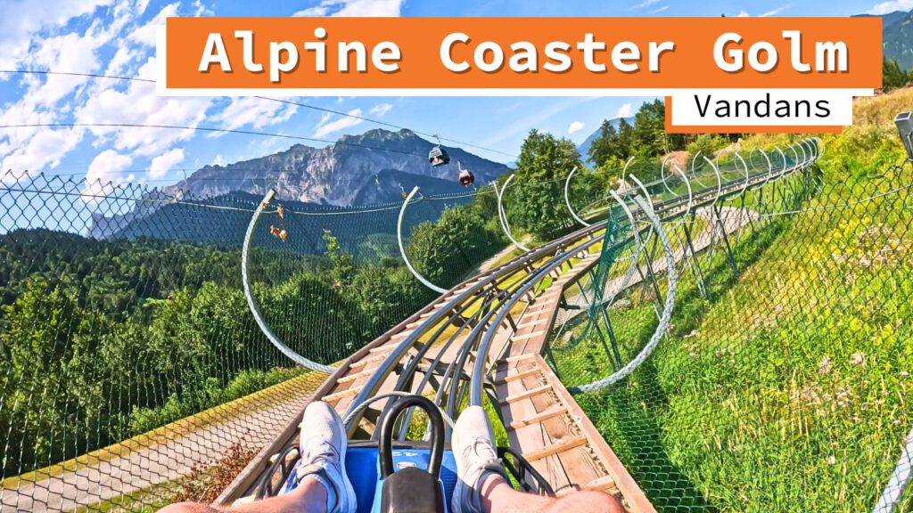 Blick aus der Perspektive eines Fahrgasts auf der Strecke des Alpine Coasters Golm in Vandans. Die Schienen winden sich durch grüne Landschaft und sind von Sicherheitszäunen umgeben, mit einem Bergpanorama im Hintergrund und Seilbahnkabinen am Himmel.