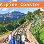 Blick aus der Perspektive eines Fahrgasts auf der Strecke des Alpine Coasters Golm in Vandans. Die Schienen winden sich durch grüne Landschaft und sind von Sicherheitszäunen umgeben, mit einem Bergpanorama im Hintergrund und Seilbahnkabinen am Himmel.