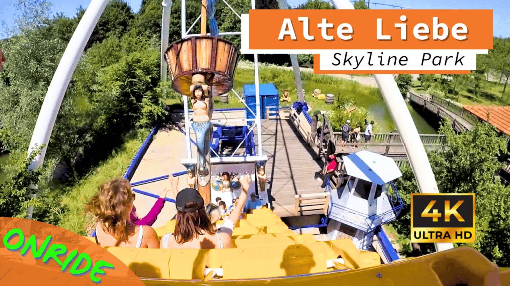 Onride-Ansicht des Schaukelschiffs „Alte Liebe“ im Skyline Park. Besucher sitzen im gelben Schiff und halten die Arme in die Luft, während das Schiff in Schwung ist. Vor ihnen ist eine geschnitzte Figur und im Hintergrund eine grüne Landschaft sowie ein hölzerner Steg mit weiteren Besuchern zu sehen. Aufnahme in 4K Ultra HD.
