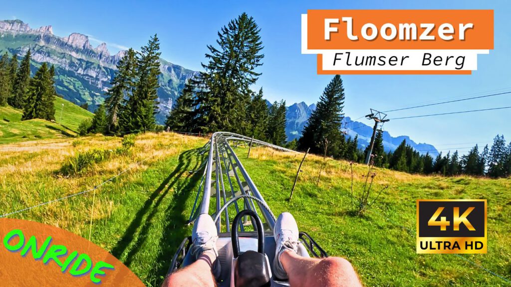 Perspektive eines Fahrgasts auf der Strecke des Floomzer Alpine Coasters am Flumser Berg. Die Schienen führen durch eine grüne Berglandschaft mit Bäumen und einem Bergpanorama im Hintergrund, unter klarem blauem Himmel.