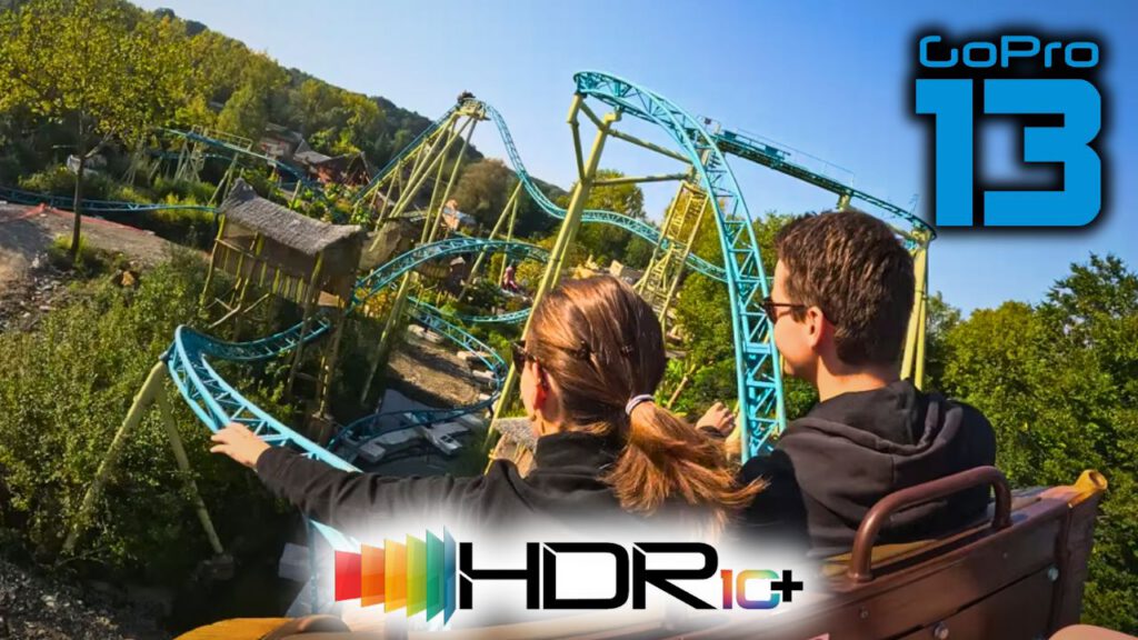 Eine Rückansicht von zwei Personen in der letzten Reihe eines Achterbahnwagens auf der Tiki Waka, einer farbenfrohen Achterbahn mit türkisfarbenen Schienen. Die Fahrt wurde mit einer GoPro HERO 13 in HDR10+ aufgenommen. Die Strecke schlängelt sich durch grüne Bäume und thematisch dekorierte Bereiche. Oben rechts ist das GoPro HERO 13 Logo und unten das HDR10+ Logo zu sehen.