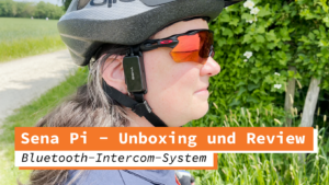 Frau mit Fahrradhelm und Sonnenbrille, die das Sena Pi Bluetooth-Intercom-System am Helm trägt. Sie steht im Freien vor einem grünen Hintergrund aus Bäumen und Wiesen. Das Intercom-Gerät ist an der Seite des Helms befestigt und ermöglicht die Kommunikation während der Fahrt.