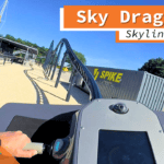 Blick aus der Onride-Perspektive des Sky Dragster im Skyline Park. Die Hände des Fahrers halten den Steuergriff des Motorrads, das auf einer schwarzen Schiene entlang einer sandfarbenen Strecke fährt. Im Hintergrund ist ein Gebäude mit der Aufschrift "Spike Racing" zu sehen. Die Aufnahme ist in 4K Ultra HD-Qualität.