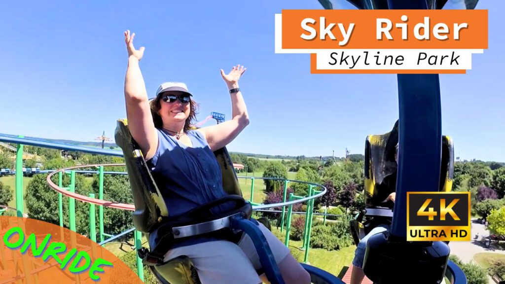 Eine Frau hebt während der Fahrt auf dem Sky Rider im Skyline Park beide Arme in die Luft. Sie sitzt in einem gelben Sitz mit Sicherheitsbügeln und trägt eine Sonnenbrille sowie einen Hut. Im Hintergrund sind grüne Bäume, Teile der Schienenstruktur und die weitläufige Parklandschaft unter einem klaren, blauen Himmel zu sehen. Auf dem Bild ist "4K Ultra HD" und "Onride" zu lesen.