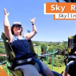 Eine Frau hebt während der Fahrt auf dem Sky Rider im Skyline Park beide Arme in die Luft. Sie sitzt in einem gelben Sitz mit Sicherheitsbügeln und trägt eine Sonnenbrille sowie einen Hut. Im Hintergrund sind grüne Bäume, Teile der Schienenstruktur und die weitläufige Parklandschaft unter einem klaren, blauen Himmel zu sehen. Auf dem Bild ist "4K Ultra HD" und "Onride" zu lesen.