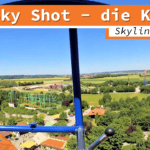 Onride-Ansicht des Fahrgeschäfts Sky Shot im Skyline Park. Die Aufnahme zeigt die Aussicht aus einer kugelförmigen Gondel in luftiger Höhe über den Park und die umliegende Landschaft. Die Gondelstruktur ist blau, und im Hintergrund sind weitere Attraktionen sowie grüne Felder und Bäume zu sehen.