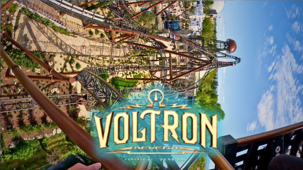Aufnahme von der Onride-Kamera der Achterbahn Voltron Nevera im Europa-Park. Die verschlungenen Schienen führen durch enge Kurven und steile Abfahrten, während eine aufwendige Landschaftsgestaltung mit Pflanzen und Steinen die Strecke umgibt. Im Hintergrund sind Gebäude und Besucher des Parks erkennbar. Zentral ist das große Voltron Nevera-Logo zu sehen, darüber spannt sich ein klarer blauer Himmel.