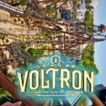 Aufnahme von der Onride-Kamera der Achterbahn Voltron Nevera im Europa-Park. Die verschlungenen Schienen führen durch enge Kurven und steile Abfahrten, während eine aufwendige Landschaftsgestaltung mit Pflanzen und Steinen die Strecke umgibt. Im Hintergrund sind Gebäude und Besucher des Parks erkennbar. Zentral ist das große Voltron Nevera-Logo zu sehen, darüber spannt sich ein klarer blauer Himmel.