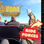 Blick von hinten auf Fahrgäste der Holzachterbahn Wodan Timburcoaster im Europa-Park, flankiert von Wikingerfiguren aus Holz, mit einem Diagramm rechts, das die vertikalen, lateralen und longitudinalen G-Kräfte der Fahrt anzeigt.